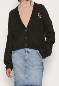 Donna che indossa un cardigan nero con bottoni e piccole ricami floreali, un maglione beige a collo alto e una gonna di jeans azzurro chiaro con tasche.