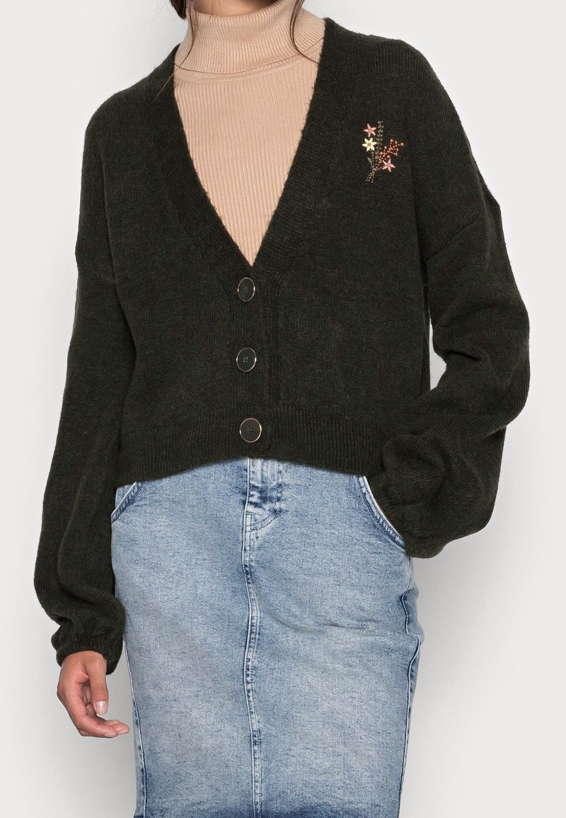 Donna che indossa un cardigan nero con bottoni e piccole ricami floreali, un maglione beige a collo alto e una gonna di jeans azzurro chiaro con tasche.