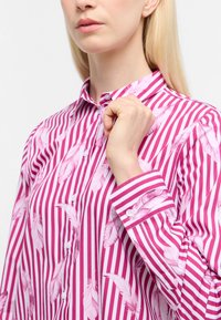Button-up skjorte med lyserøde og hvide striber, prydet med fjer-motiver. Fremstillet af letvægts stof med klassisk krave og manchetter.