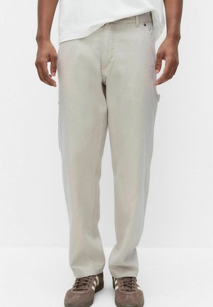 Mężczyzna ubrany w jasnobeżowe codzienne spodnie i brązowe trampki z białymi paskami, stojący na jednolitym białym tle.