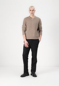 Beige knitted sweater with a textured pattern and a V-neck, paired with black trousers. Model stands in a neutral background.