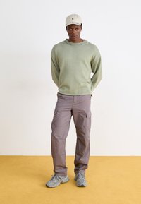 Lys grønn genser med ribbet hals, grå cargo-bukser og grå sneakers, toppet med en beige caps, mot en gul gulv.