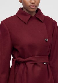 Femme portant un manteau cache-cœur bordeaux avec un grand col et des boutons argentés, noué à la taille avec une ceinture en tissu assorti.