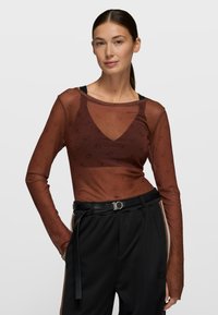 Top marrón de manga larga y transparente con un patrón sutil, combinado con pantalones negros y un cinturón negro texturizado con un detalle de hebilla redonda.