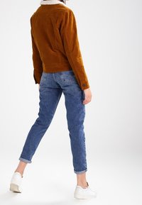 Bruine ribfluwelen jas met een witte sherpa kraag, gecombineerd met een blauwe spijkerbroek en witte sneakers, gezien van achteren. Casual pasvorm en omgeslagen mouwen.