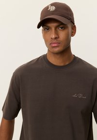 Καφέ βαμβακερό T-shirt με στρογγυλή λαιμόκοψη, στολισμένο με ένα διακριτικό κεντητό λογότυπο σε ανοιχτότερο καφέ. Φορεμένο με καπέλο μπέιζμπολ στο ίδιο καφέ χρώμα.