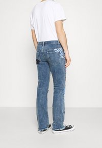 Jeans de mezclilla azul con dobladillo deshilachado, detalles de patchwork y varios acentos de tela en gris y azul. Llevados con una camiseta blanca sencilla y zapatillas negras.