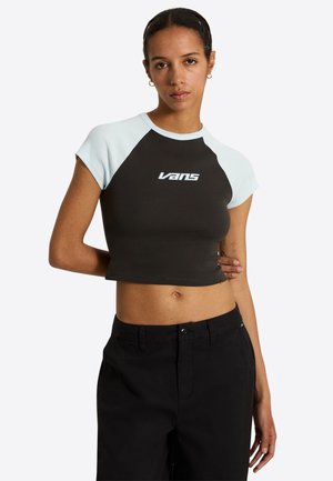Nainen pukeutunut mustaan lyhennettyyn Vans-paitaan, jossa valkoiset hihat, seisoo käsi selän takana ja päällään mustat housut.