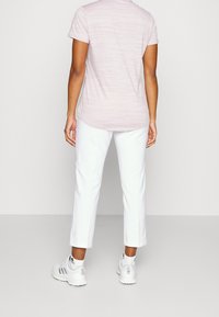 Person med ljusrosa kortärmad skjorta, vita korta byxor, vita Adidas-sneakers, stående mot en enfärgad ljus bakgrund, bakvy.