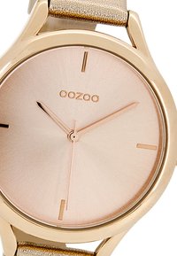 Montre dorée avec un cadran rond en or rose, des index minimalistes et un bracelet en tissu de couleur claire. La marque "OOZOO" est affichée.