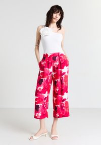 Haut blanc à une épaule avec un détail de fleur en tissu, associé à un pantalon ample à motif floral rouge sur fond blanc.