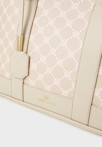 Sac à main beige en tissu à motifs présentant des designs circulaires blancs. Accents en cuir lisses et logo doré sur la base. Détail de fermeture éclair.