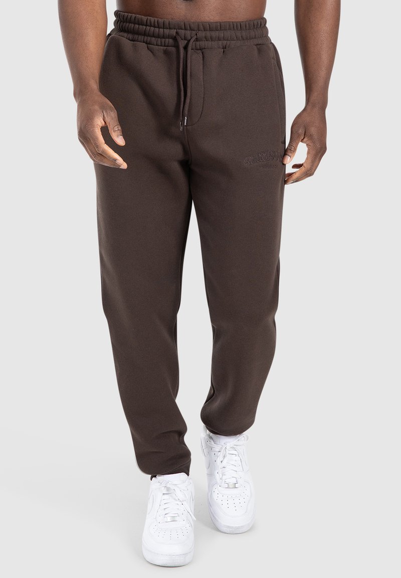 Mann trägt dunkle braune Sweatpants mit elastischem Bund und weiße Sneakers, steht vor einem einfarbigen Hintergrund.