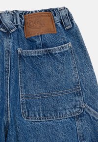 Denim jeans med blå tekstur, med en brun lærmerkel ved livbåndet og en baklomme. Synlig sømdetalj.