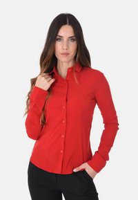 Camicia rossa a maniche lunghe con chiusura a bottoni, colletto e design slim. Presenta un tessuto morbido e un orlo arrotondato. Indossata con pantaloni neri.