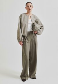 Grå kort jakke med rund halslinning og ballongsleeves, parret med løse olivengrønne bukser. Holder en mønstret veske.