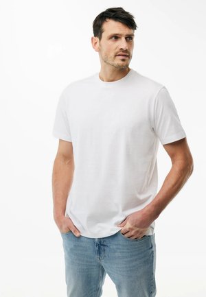 Hombre de pie con las manos en los bolsillos, vistiendo una camiseta blanca de manga corta lisa y jeans azul claro, mirando hacia su izquierda.