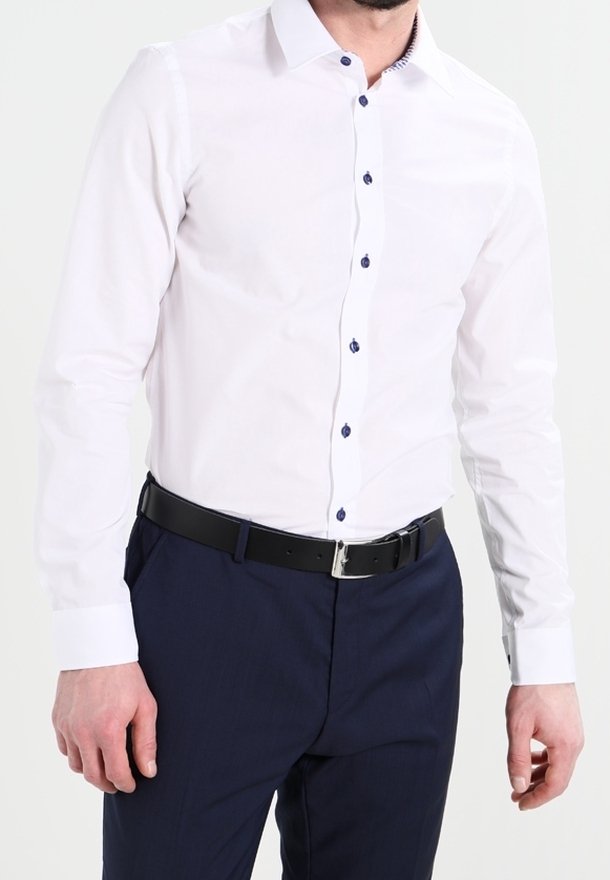 Chemise blanche à boutons bleus, manches longues et col, portée avec un pantalon bleu marine et une ceinture noire avec une boucle argentée.