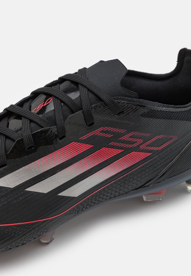 Botín de fútbol negro con una superficie texturizada, que presenta detalles en plateado metálico y rojo, un diseño elegante y un sistema de cordones tradicional.