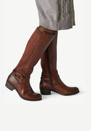 Bottes en cuir marron montant jusqu’aux genoux avec des talons bas et des brides décoratives à boucle, portées avec des collants foncés et une jupe en tricot grise.