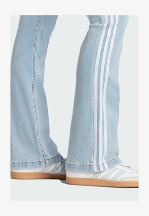 Helle blaue, ausgestellte Jeans mit weißen Seitestreifen, kombiniert mit weißen Sneakers mit grauen Akzenten und einer Gummisohle. Sichtbare Nähte am Saum.