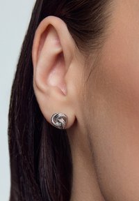 Srebrne kolczyki w kształcie skręconego węzła z misternymi detalami. Umieszczone na zbliżeniu ucha, podkreślając fakturę i kształt.