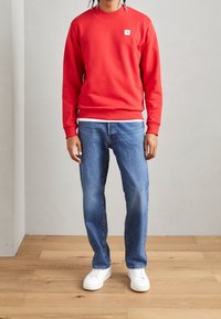 Sudadera roja con cuello redondo, puños y dobladillo acanalados; combinada con vaqueros azules y zapatillas blancas. Presenta un pequeño logo en el pecho.