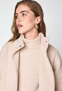 Donna con capelli castano chiaro, che indossa un maglione beige a collo alto, un cappotto abbinato e grandi orecchini a cerchio in oro texturizzato, mentre guarda verso sinistra.