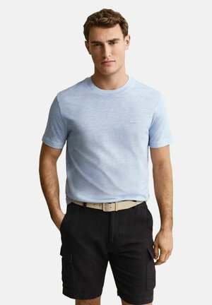 Homme portant un t-shirt bleu clair à col rond, une ceinture beige et un short cargo noir, les mains dans et près des poches, sur fond blanc.