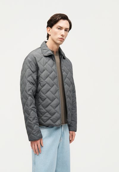 Νεαρός άνδρας με γκρι ζακέτα με φερμουάρ και ραφές, καφέ πουλόβερ και ανοιχτό μπλε τζιν, στέκεται μπροστά σε ένα απλό φωτεινό φόντο.