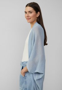 Lichtblauwe gebreide kimono cardigan met open voorkant, lange mouwen en een gestructureerde stof; gecombineerd met een witte top en denim shorts.