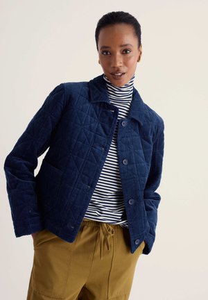 Femme portant une veste matelassée bleu marine, un col roulé rayé et un pantalon moutarde avec cordon, sur un fond clair uni.