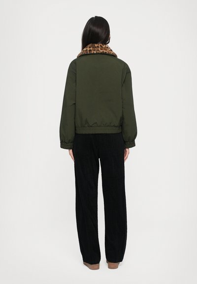 Olivinės spalvos bomber tipo striukė su leopardų rašto apykakle, elastingais rankogaliais ir liemeniu, derinama su juodais veliūro keliais.