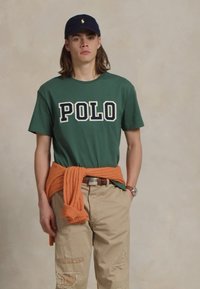 Jaunas žmogus, vilkintis žalią "POLO" marškinėlį, mėlyną kepurę, kreminės spalvos nuvalytus kelnes ir oranžinį megztinį, surištą aplink liemenį, stovi prieš paprastą foną.