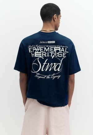 Férfi tengerészkék pólóban, melynek hátulján fehér felirat olvasható: „Ephemeral Heritage Stwd Since 1991 Respect the Legacy”, világos bézs nadrággal párosítva.