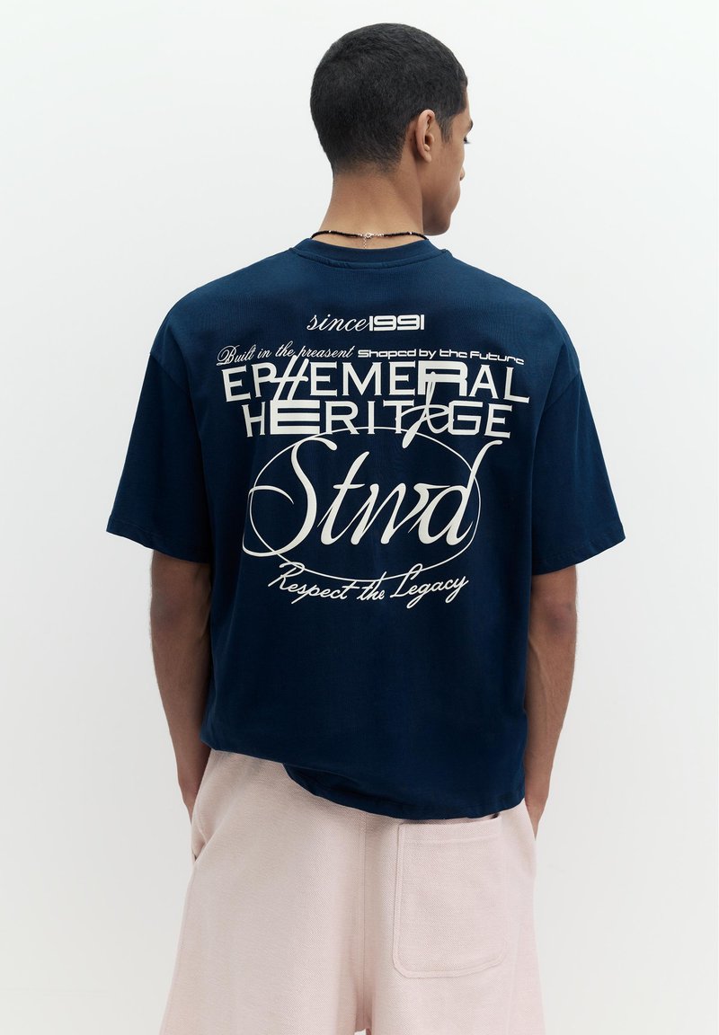Homme portant un t-shirt bleu marine avec un texte blanc au dos disant "Ephemeral Heritage Stwd Since 1991 Respect the Legacy", associé à un pantalon beige clair.