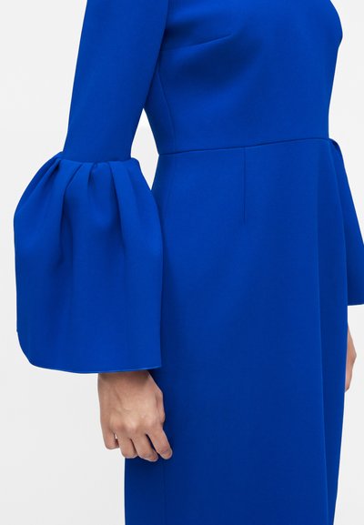 Φωτεινό μπλε φόρεμα με εφαρμοστή μέση και δραματικά φουσκωτά μακριά μανίκια που τελειώνουν σε φαρδιές μανσέτες στον μπράτσο του μοντέλου.