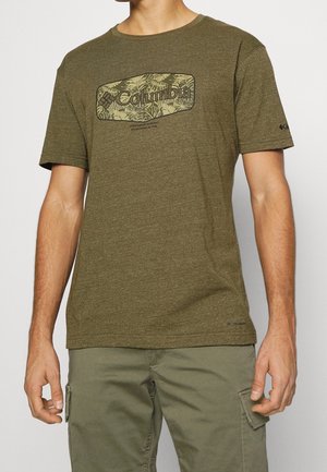 Mann trägt olivgrünes Columbia-T-Shirt mit Blattmuster-Logo und passende Cargohosen, steht vor schlichtem Hintergrund.