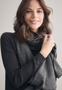 Grå uldscarf draperet løst omkring halsen, kombineret med en sort langærmet skjorte. Blød tekstur og afslappet stil.