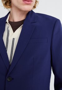 Mornarica blazer iz gladke tkanine z enim gumbom, zareznim ovratnikom in žepom na prsih. Nosi se čez smetanasto pulover s črnimi vzorci.