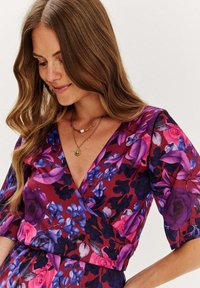 Czerwona sukienka w kwiaty, z fioletowymi i niebieskimi wzorami, z dekoltem w kształcie litery V i krótkimi rękawami, wykonana z lekkiego materiału.