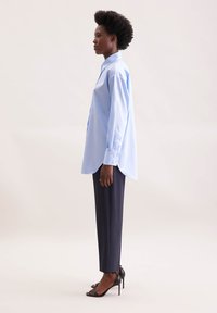 Camicia azzurro chiaro a maniche lunghe con dettagli di bottoni e orlo arrotondato, abbinata a pantaloni scuri sartoriali e sandali neri con tacco alto.