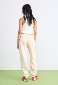 Balta cropped top, krēmkrāsas bikses ar elastīgu jostasvietu un gaiši rozā sporta apaviem. Fons ir ar zaļu grīdu un baltu sienu.