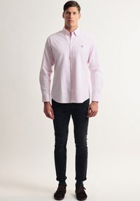 Camisa de botones de color rosa claro con rayas blancas, de manga larga y un pequeño logo en azul marino en el pecho, combinada con pantalones oscuros ajustados.