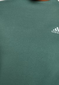 Camiseta verde de cuello redondo hecha de tela suave, con un pequeño logo blanco de Adidas en el pecho. Textura suave con elementos de diseño mínimos.