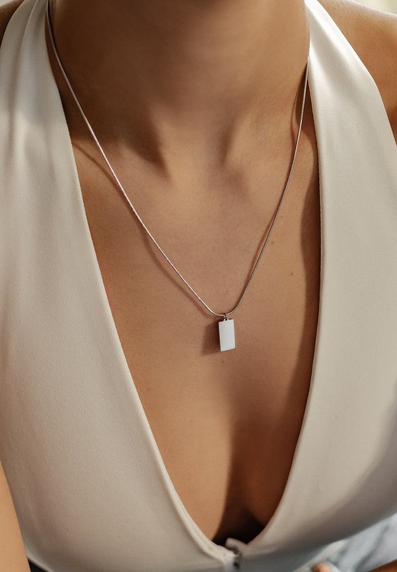 Collar de plata con un colgante rectangular blanco, llevado sobre una blusa de color claro con un escote en V profundo. Diseño minimalista y elegante.