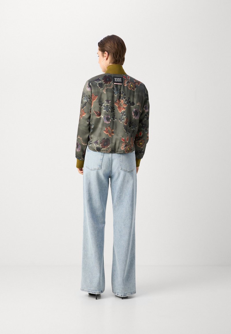 Personne avec les cheveux courts portant une veste bomber à motif floral et un jean large bleu clair, se tenant contre un fond blanc uni.