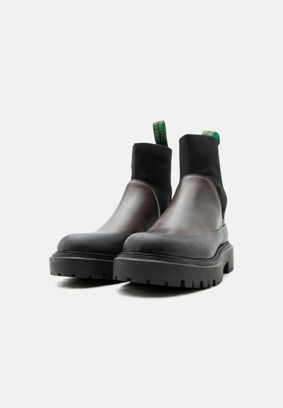 Ζευγάρι μαύρα και σκούρα καφέ μποτάκια μέχρι τον αστράγαλο με χοντρές σόλες και πράσινες υφασμάτινες θηλιές, σε λευκό φόντο.