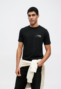 Czarny bawełniany t-shirt z krótkimi rękawami i okrągłym dekoltem. Na lewej piersi znajduje się biały nadruk tekstowy. Noszony z kremowym swetrem zawiązanym w talii.