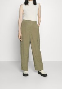 Pantaloni cargo khaki con gambe larghe, dotati di due tasche laterali, realizzati in tessuto liscio. Abbinati a un top aderente senza maniche color crema e a scarpe slip-on.